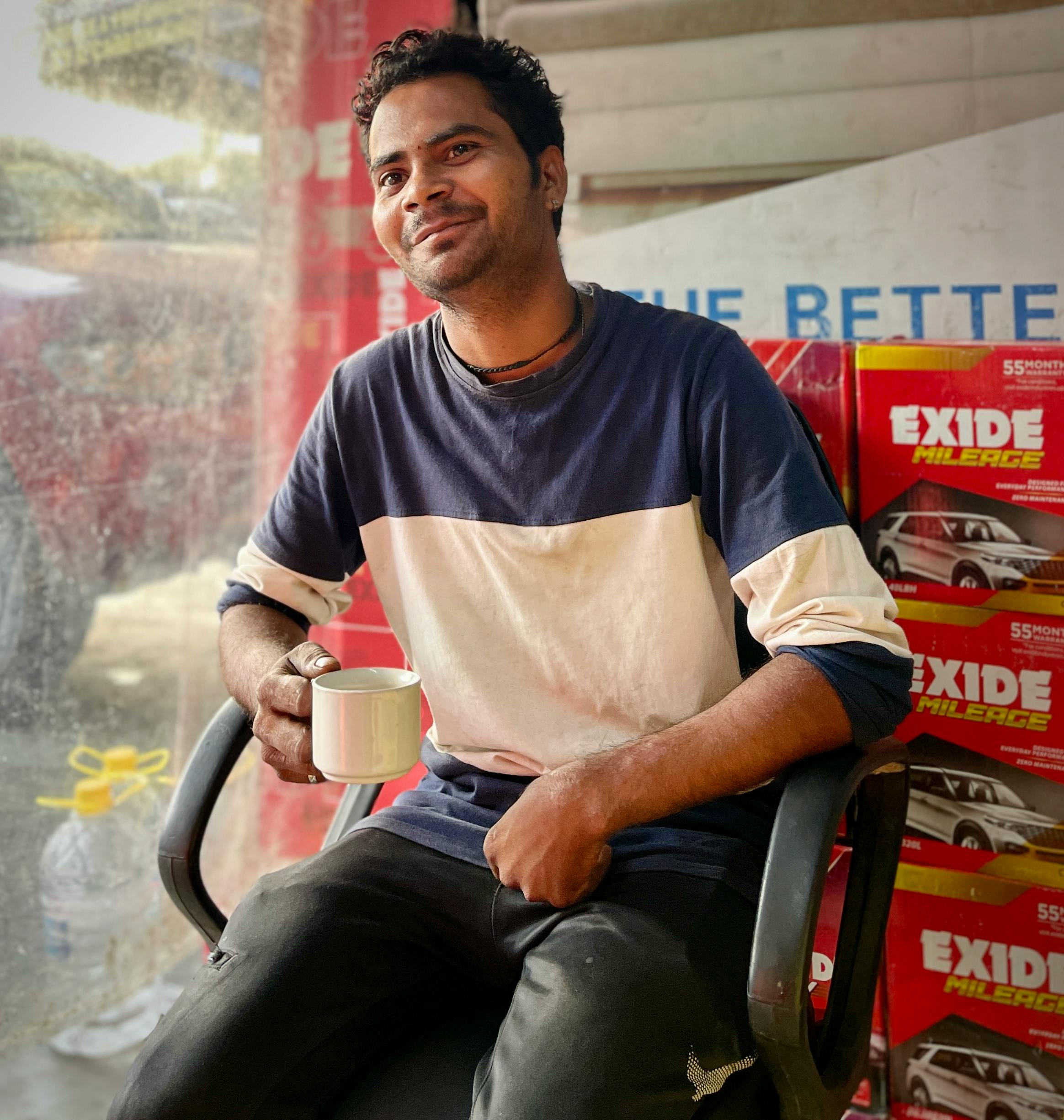 Smiling man enjoying coffee at a car battery shop, sitting by the window.