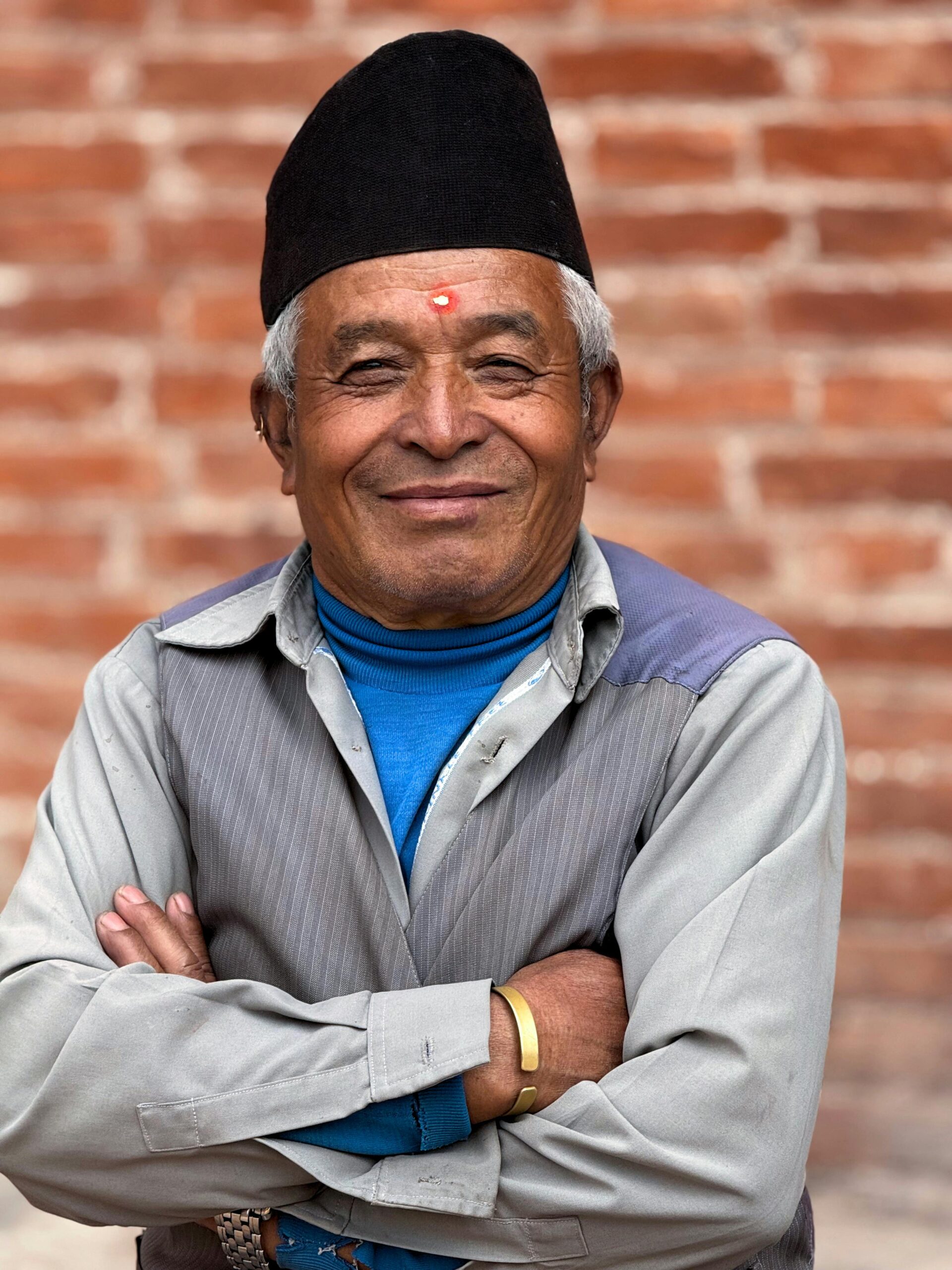 Portrait of an elderly man with a serene expression in Bhaktapur, Nepal, wearing traditional clothing against a brick wall.